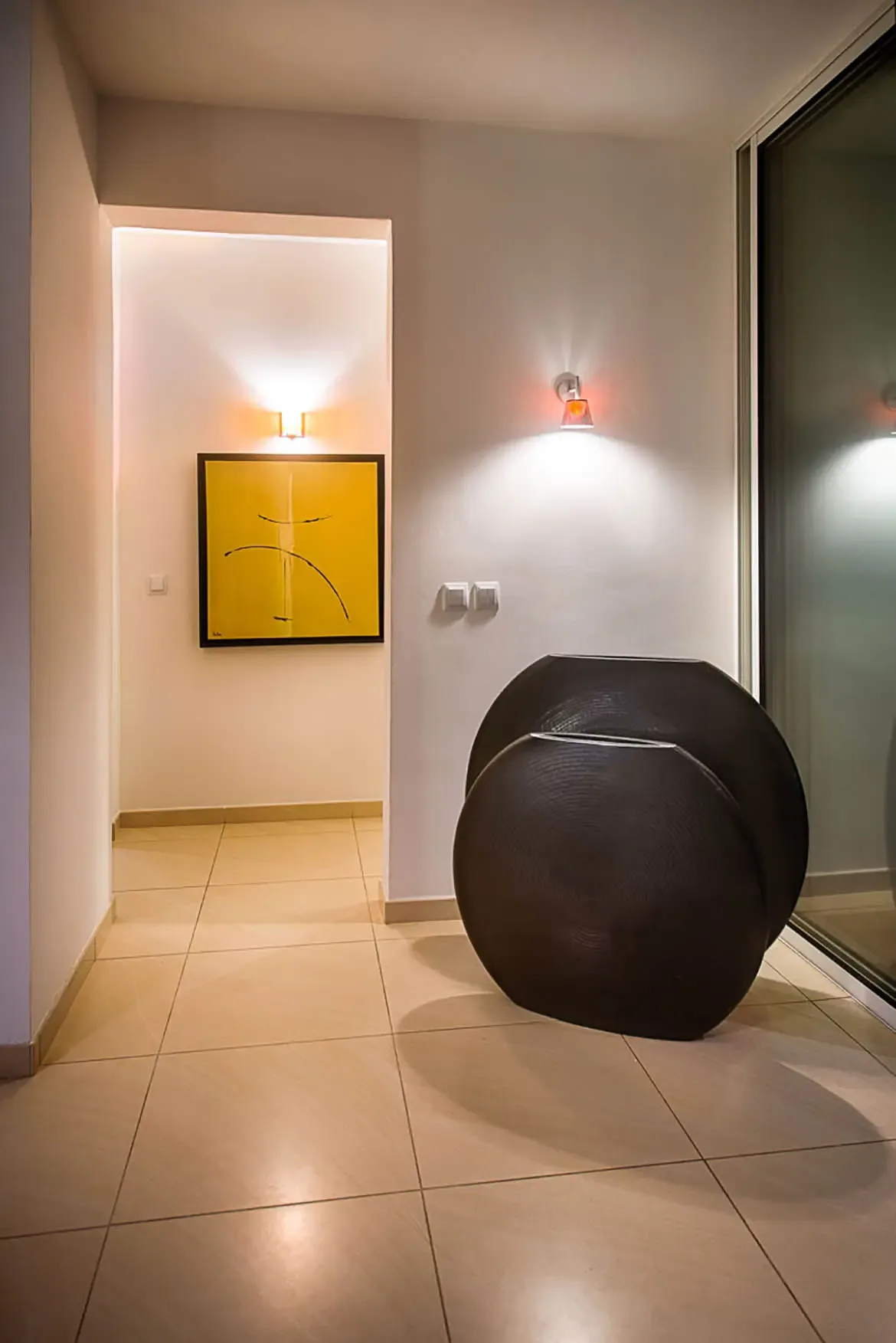 A modern hallway featuring yellow abstract painting, two large dark circular sculptural vases, and white wall with small sconce.