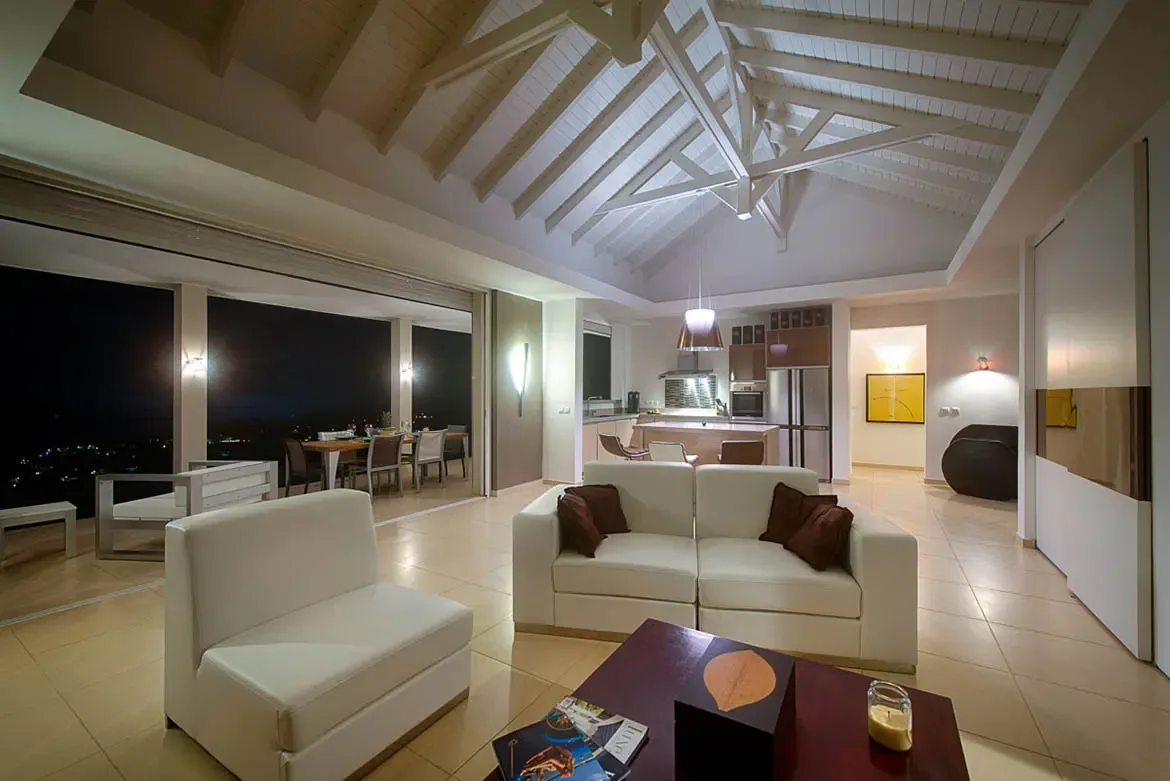 A wide-angle evening shot of open-plan living area with vaulted white beam ceilings, white leather sofa, kitchen with stainless steel appliances, and covered terrace overlooking city lights.