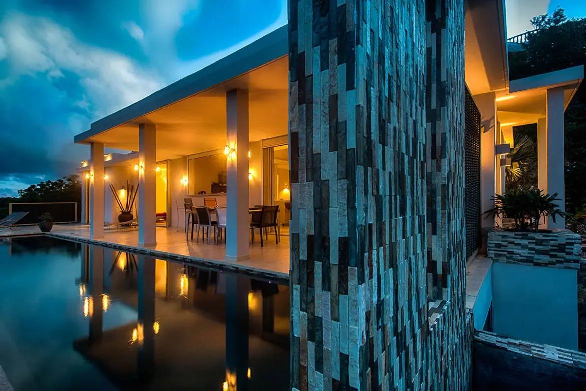 A wide-angle evening shot of modern villa with lit terrace, dining furniture reflected in large swimming pool, textured stone feature wall, and dramatic twilight sky.