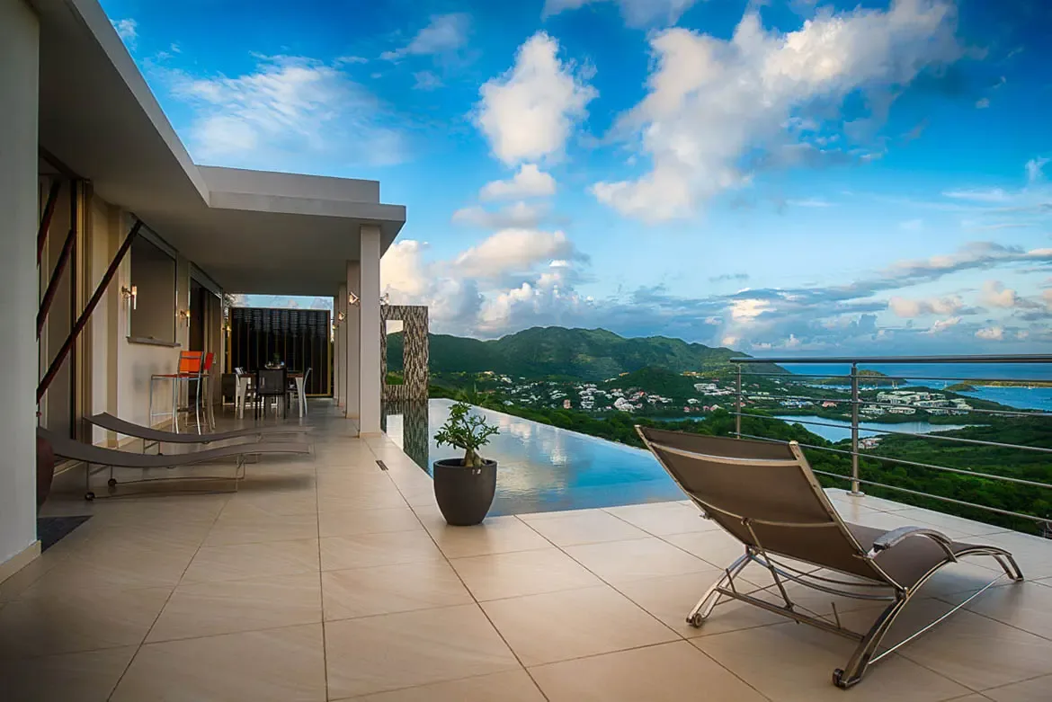 A wide-angle shot of luxurious outdoor terrace with infinity pool, modern lounge chairs, dining area, bar seating, and breathtaking panoramic view of lush hills, coastal town, and ocean.