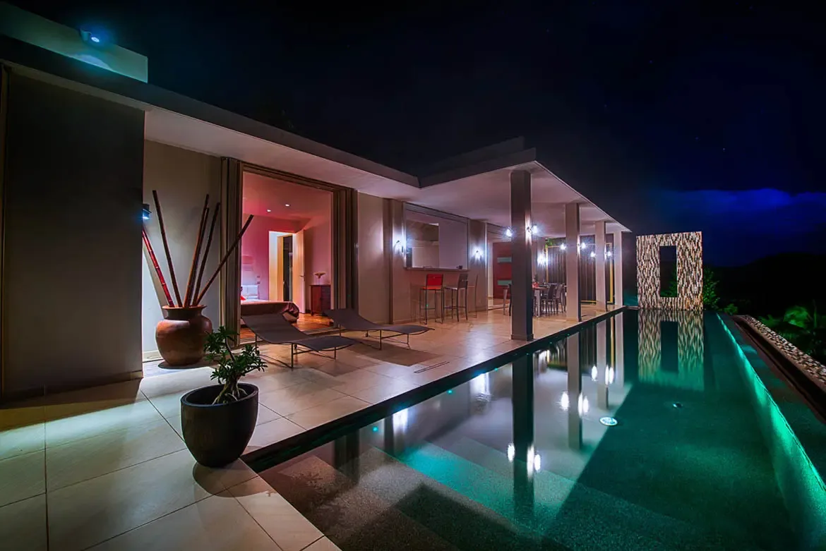 A wide-angle evening shot featuring a long infinity pool with green illumination, reflecting the villa's architecture with open-plan dining and bedroom views.