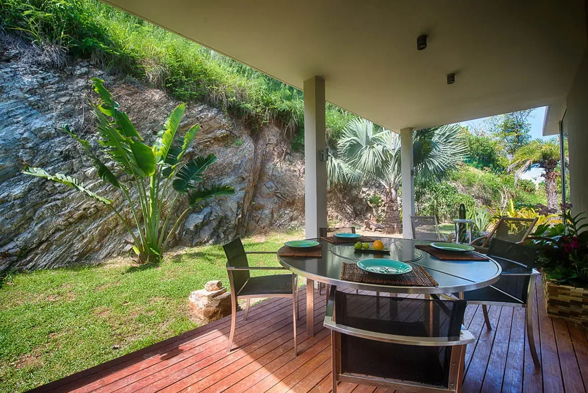 A covered outdoor dining area on wooden deck with modern round table for four, overlooking manicured lawn and striking natural rock cliff.