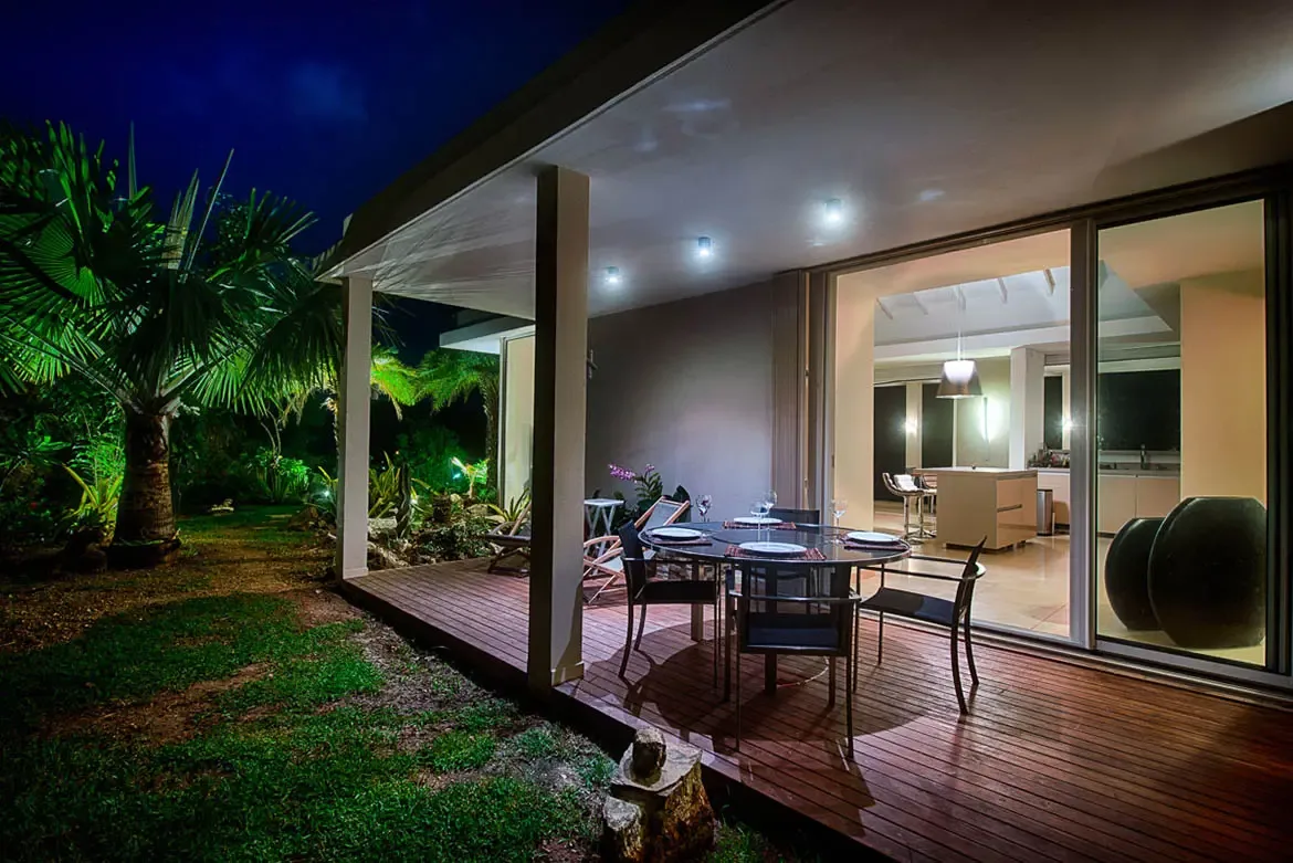 A night-time view of wooden deck with round outdoor dining set, illuminated by recessed ceiling lights and green garden spotlights on tropical palm trees.