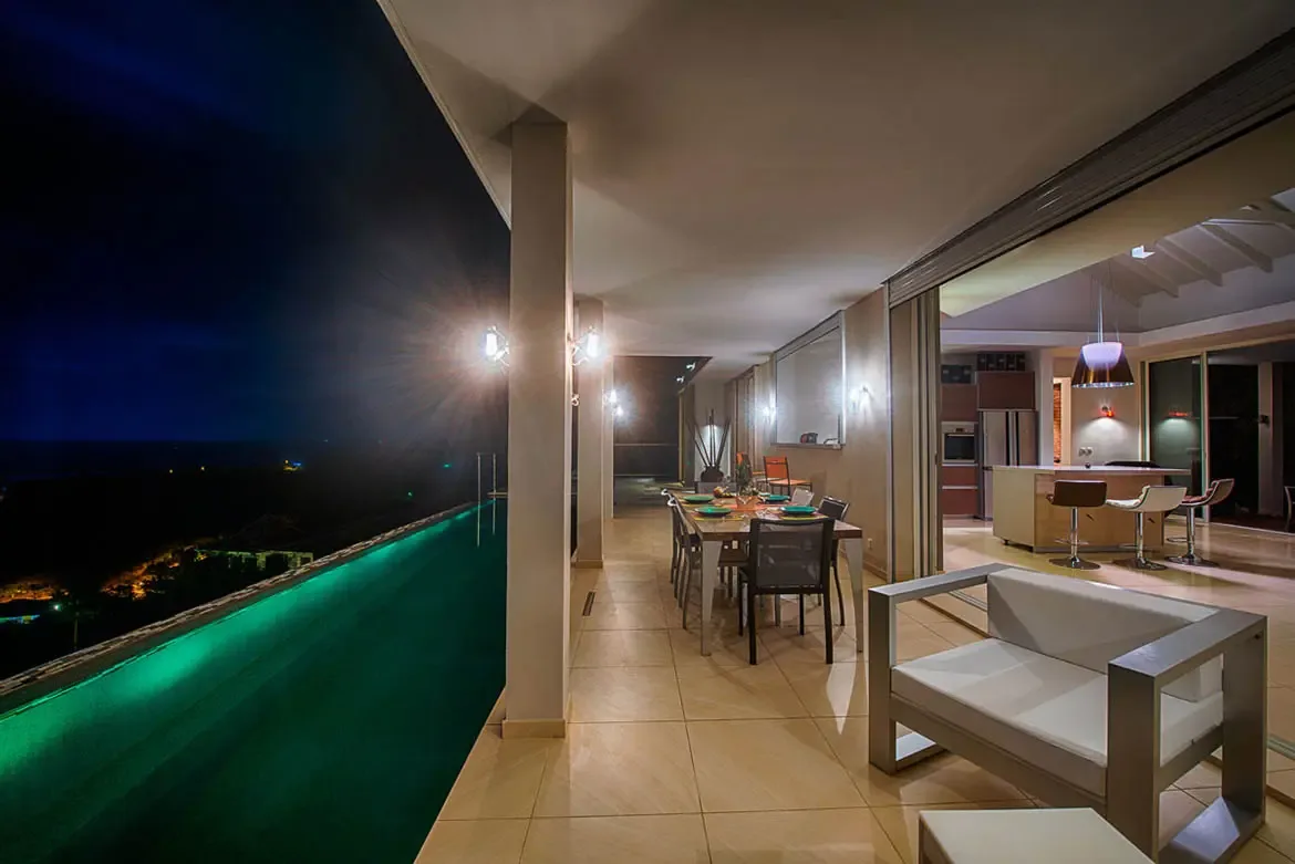 A wide-angle night shot of outdoor terrace with illuminated infinity pool, modern dining set, lounge chair, and indoor open-plan kitchen with city lights in distance.