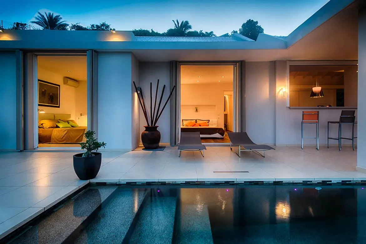 A high-end twilight view of the terrace and infinity pool with lounge chairs and two bedrooms visible through large sliding glass doors.