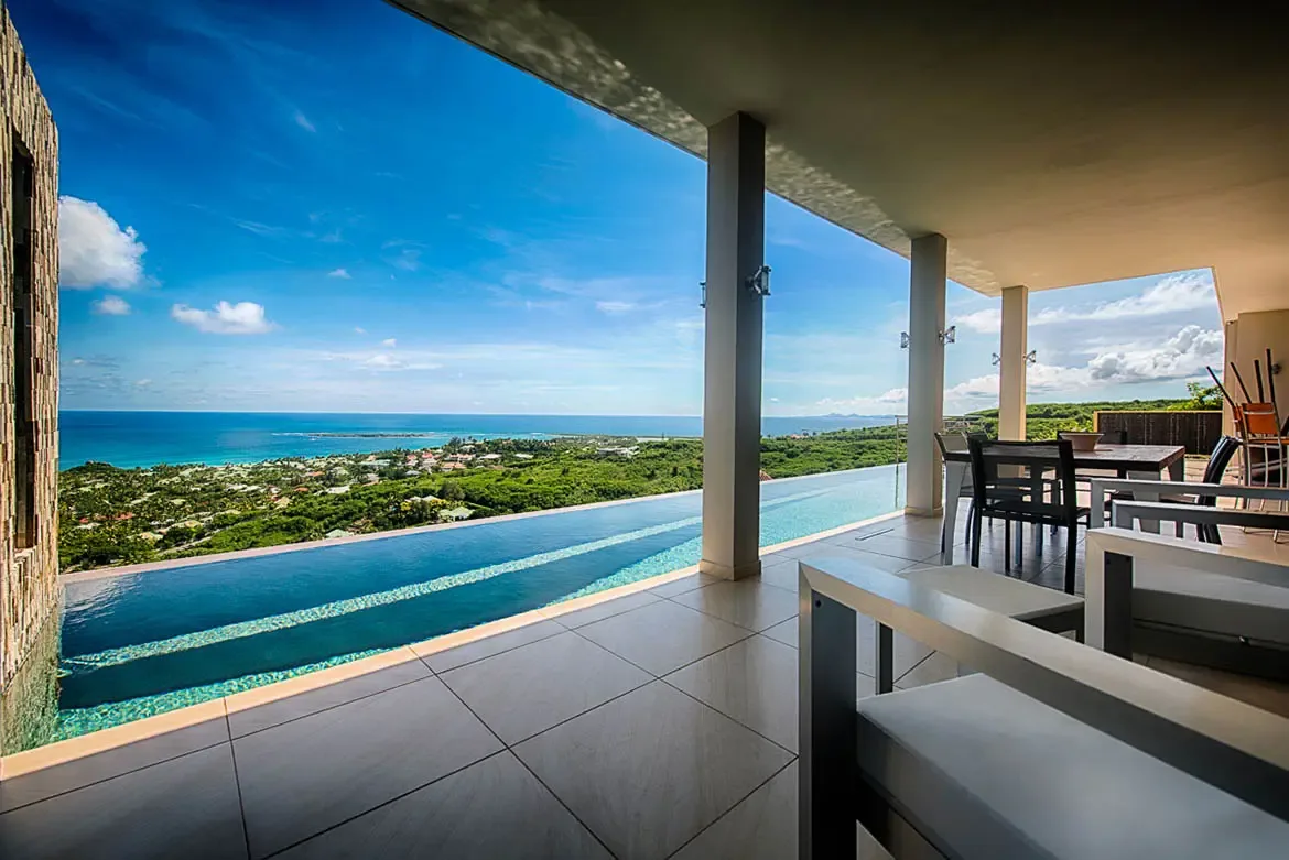 An expansive wide-angle shot from covered patio with tiered infinity pool, overlooking lush green landscape and turquoise ocean with modern outdoor lounge and dining furniture.