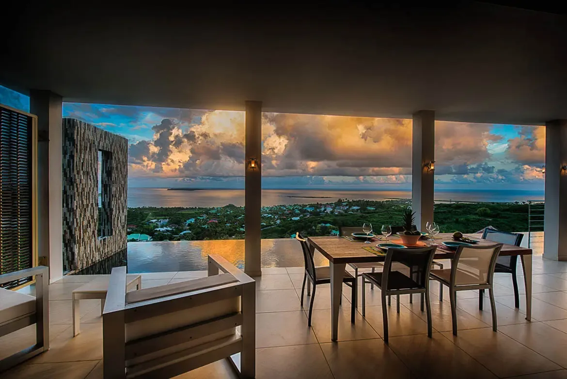 An expansive covered terrace with large wooden dining table, overlooking infinity pool blending into panoramic view of coastal town and ocean under dramatic sunset.