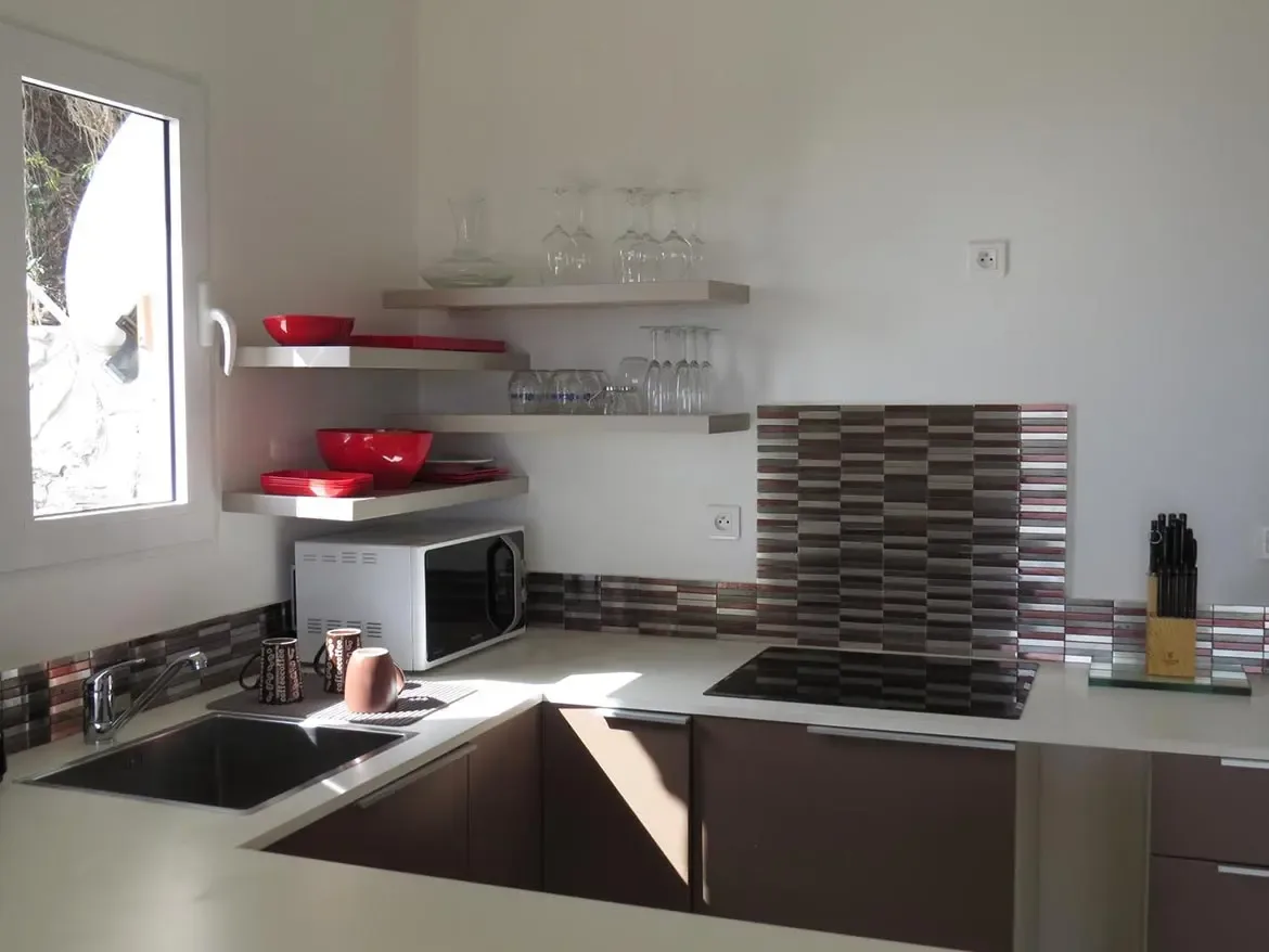 A compact modern kitchenette with white countertops, dark brown cabinetry, open floating shelves, induction stovetop, and decorative horizontal-patterned tile backsplash.