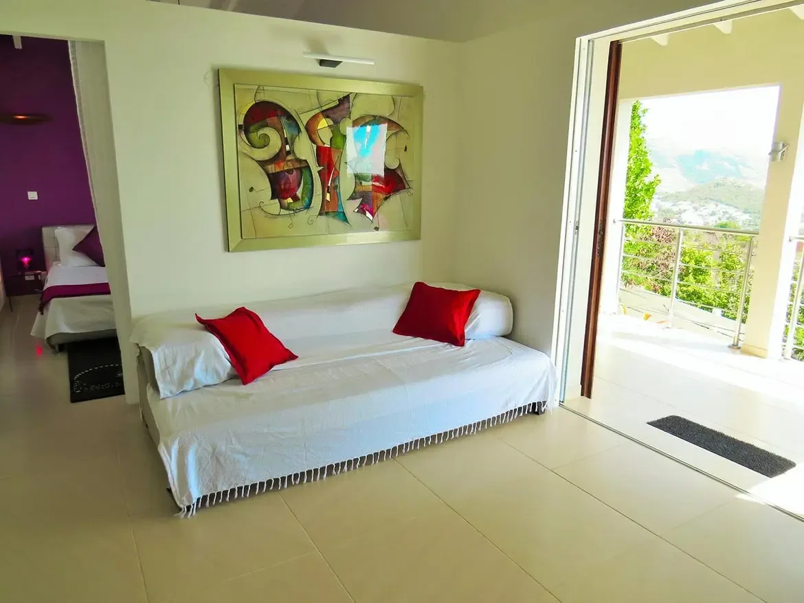 A bright living space with white fringed sofa, red accent pillows, large abstract modern art in a gold frame, and open flow to a balcony with mountain views.