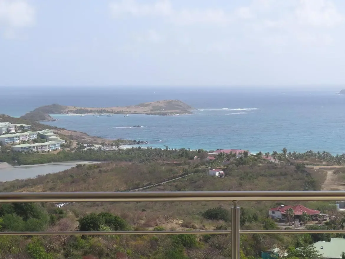 An elevated panoramic view from a balcony overlooking tropical coastline, salt pond, white resort buildings, and a distant island.