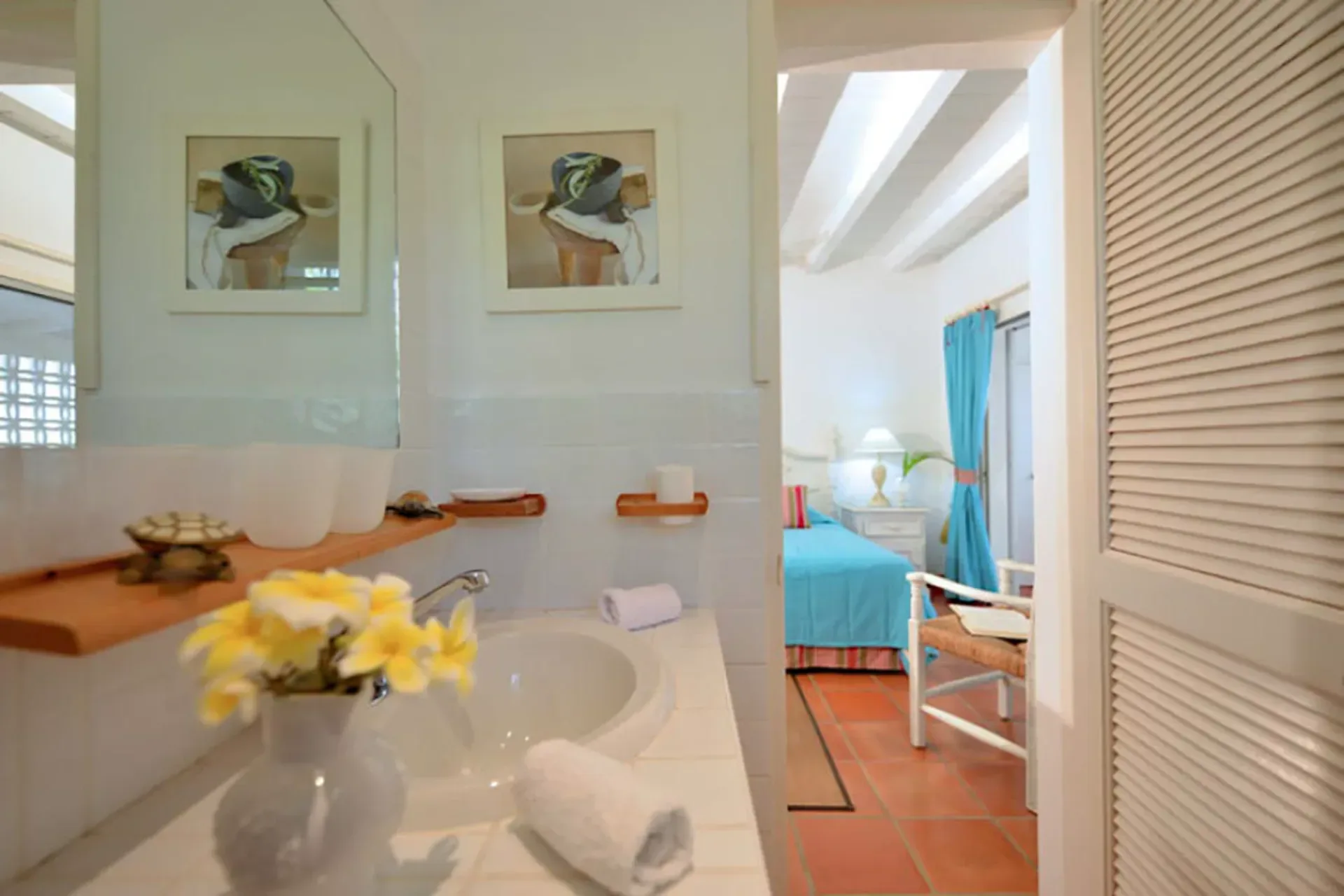 A bright bathroom with fresh flowers, opening to a bedroom with blue linens and classic Caribbean louvered doors.