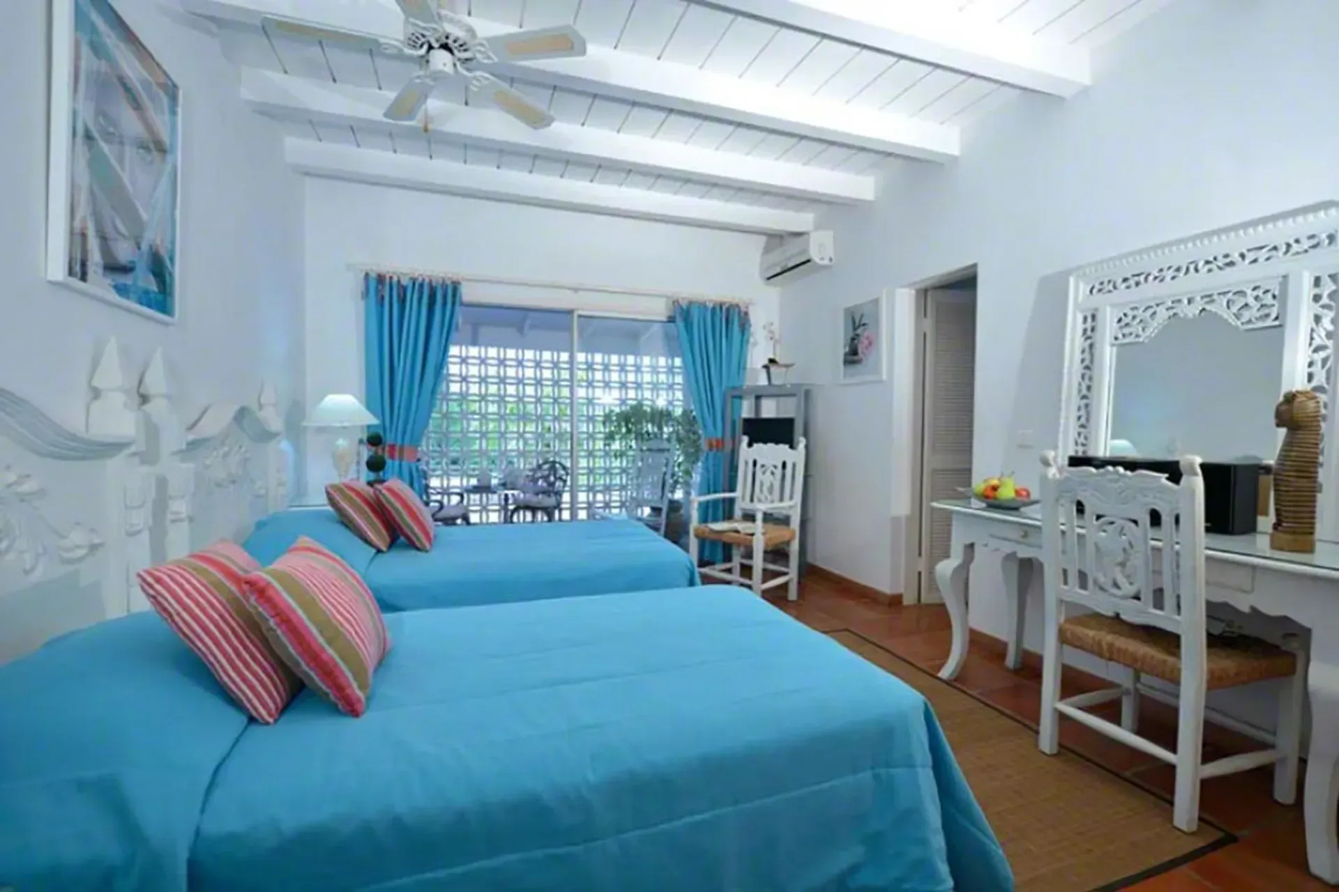 A bright twin bedroom with turquoise linens, white carved headboard, and terrace access.