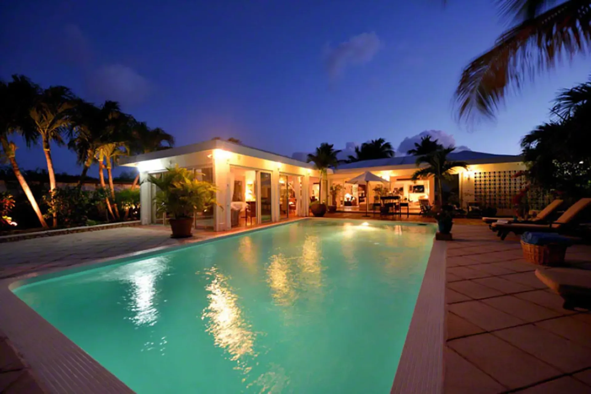 Your illuminated pool at night with modern villa architecture and tropical gardens.